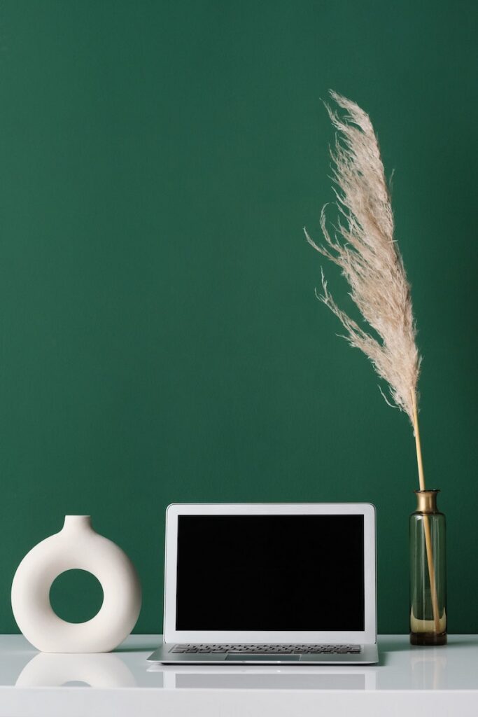 an open laptop on a clear desk surface with one piece of pampas grass sitting to the right of the laptop and an empty vase on the left
