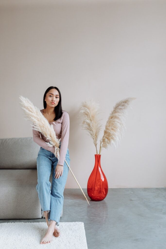 A woman leaning on the arm of a couch with a piece of pampas grass as tall as herself beside a large clear red glass vase on the floor which holds two pieces of pampas grass of similar size